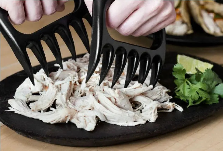 Black meat shredder claws being used to shred chicken on a plate with herbs and lime.